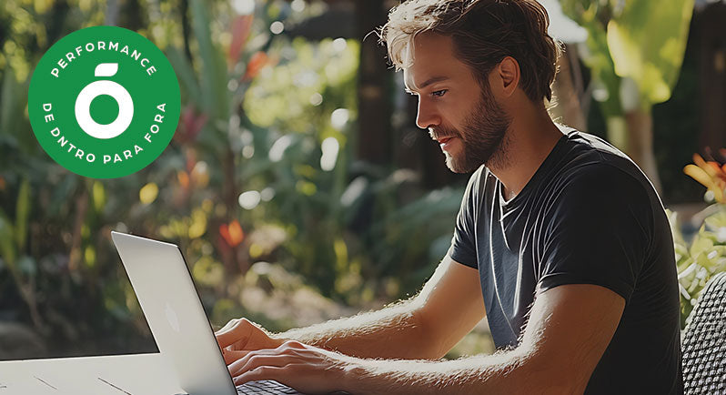 Homem concentrado trabalhando em um notebook ao ar livre, cercado por vegetação, com o selo verde “Performance de DNTRO para Fora” no canto da imagem. Representa equilíbrio entre produtividade, bem-estar e estilo de vida saudável com suporte natural.