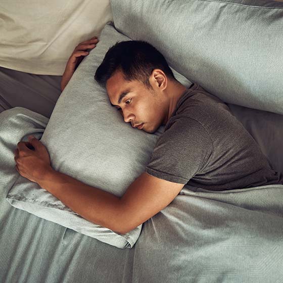 Homem de feições orientais deitado de lado na cama, com olhos abertos, abraçando travesseiro