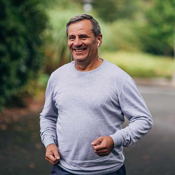 Foto de homem com cabelos grisalhos correndo ao ar livre, com árvores ao redor. Ele está sorrindo, usa camiseta de manga longa cinza e está com fones de ouvido