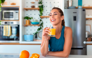 Foto de mulher branca na cozinha tomando suceo de laranja. Ela veste camiseta regata azul e está com os cabelos presos