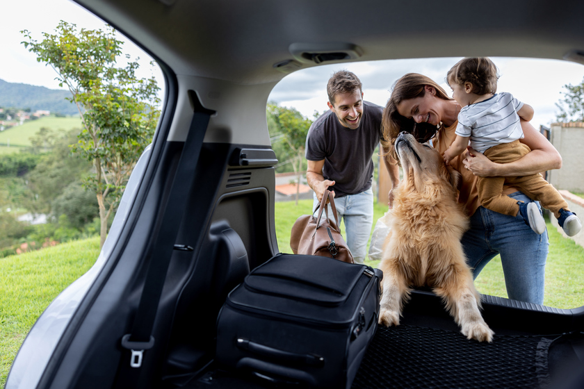 Foto de homem, mulher e criança colocando malas no porta-malas do carro. Há também um cachorro.