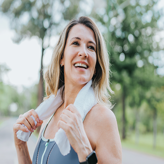 Foto de mulher branca, cabelos loiros, sorrindo no parque, com roupa de fazer exercício. Ela tem uma toalha em volta do pescoço